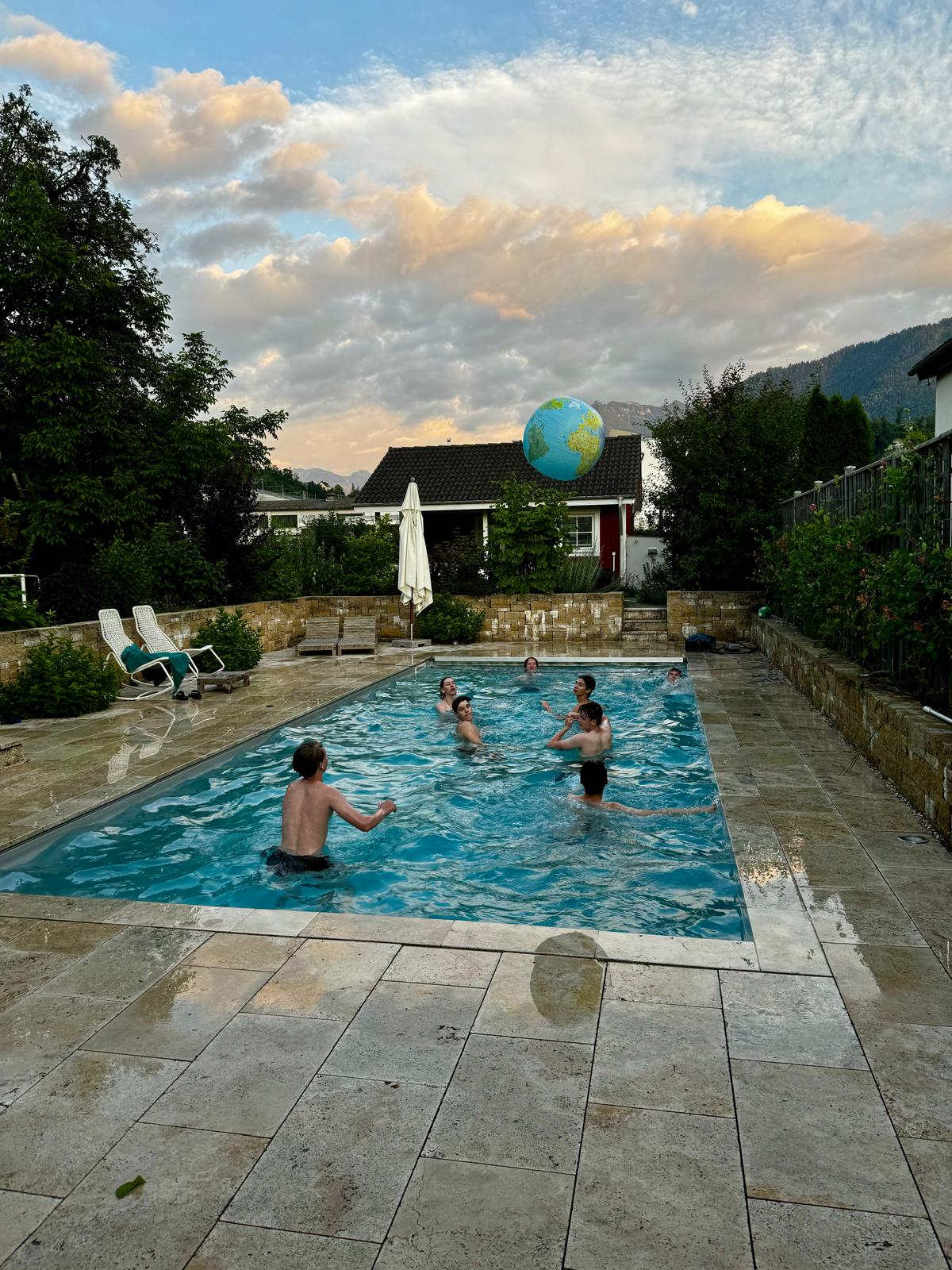 Mehrere Jugendliche schwimmen abends in einem Pool, ein aufblasbarer Globus schwebt über dem Wasser